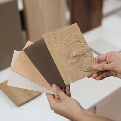 Eine Person hält Muster für Tischplatten mit verschiedenen Oberflächen, darunter Holzoptik und neutrale Farben. Im Hintergrund sind weitere größere Musterplatten sichtbar.