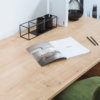 Eine Schreibtischplatte mit der Oberfläche 'Chalet Eiche' in heller Holzoptik. Darauf ein geöffnetes Magazin, zwei Stifte und ein schwarzer Organizer. Im Hintergrund eine dekorative Glasvase und weitere minimalistische Details.