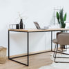 Ein minimalistischer Schreibtisch mit einer Tischplatte in Holzoptik und einem schwarzen Metallrahmen. Darauf Deko-Elemente und ein Laptop. Ergänzt wird der Raum durch einen beigen Stuhl, Pflanzen und geflochtene Körbe auf hellem Holzboden.
