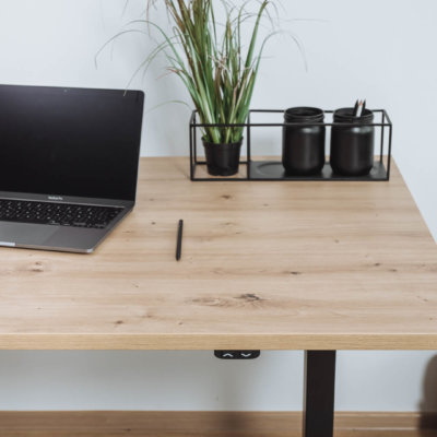 Ein Schreibtisch nach Maß mit der Oberfläche 'Atram Eiche' in heller Holzoptik. Darauf stehen ein Laptop, ein Stift und ein Organizer mit schwarzen Dekoelementen. Im Hintergrund eine grüne Pflanze.