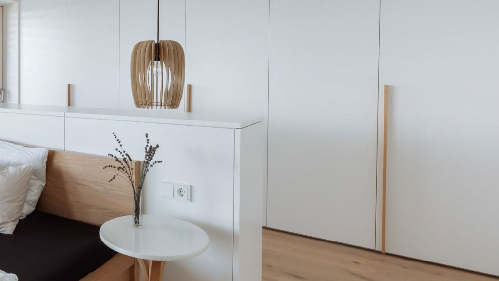 Minimalistisches Schlafzimmer mit einer Wand und Möbeln in der Oberfläche 'Topweiss matt'. Ein Kopfteil aus Holz integriert sich harmonisch mit dem weißen Design. Auf einem kleinen weißen Beistelltisch steht eine Vase mit getrockneten Lavendelzweigen. Eine dekorative Holzlampe hängt an der Wand und setzt einen natürlichen Akzent.