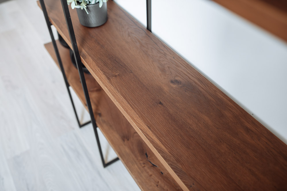 Detailaufnahme eines Regals mit rustikaler Oberfläche 'Eiche Crack'. Die dunkle Holzmaserung und natürliche Risse verleihen der Oberfläche eine authentische, charaktervolle Optik. Das Regal wird von einem schwarzen, schlanken Metallrahmen getragen, der einen modernen, industriellen Stil unterstreicht. Der Boden besteht aus hellem Holz, das einen dezenten Kontrast zur dunklen Eichenoberfläche bietet.