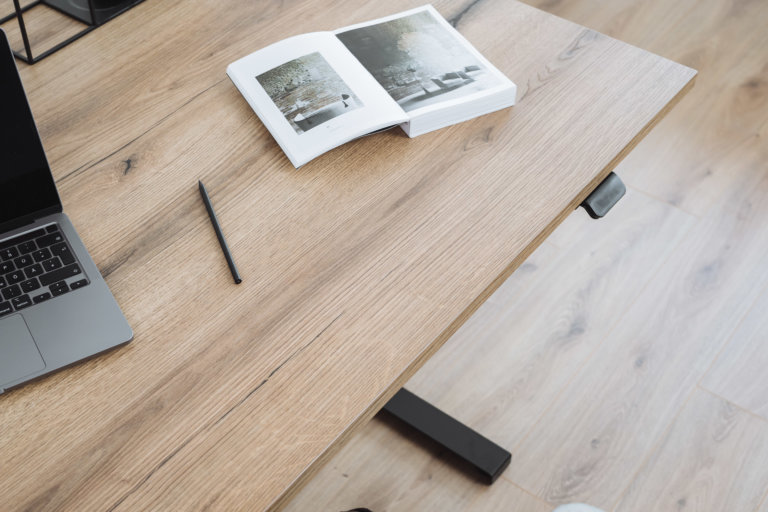 Detailaufnahme einer Tischplatte in heller Holzoptik mit natürlicher Maserung. Darauf ein Laptop, ein geöffnetes Buch und ein Stift.