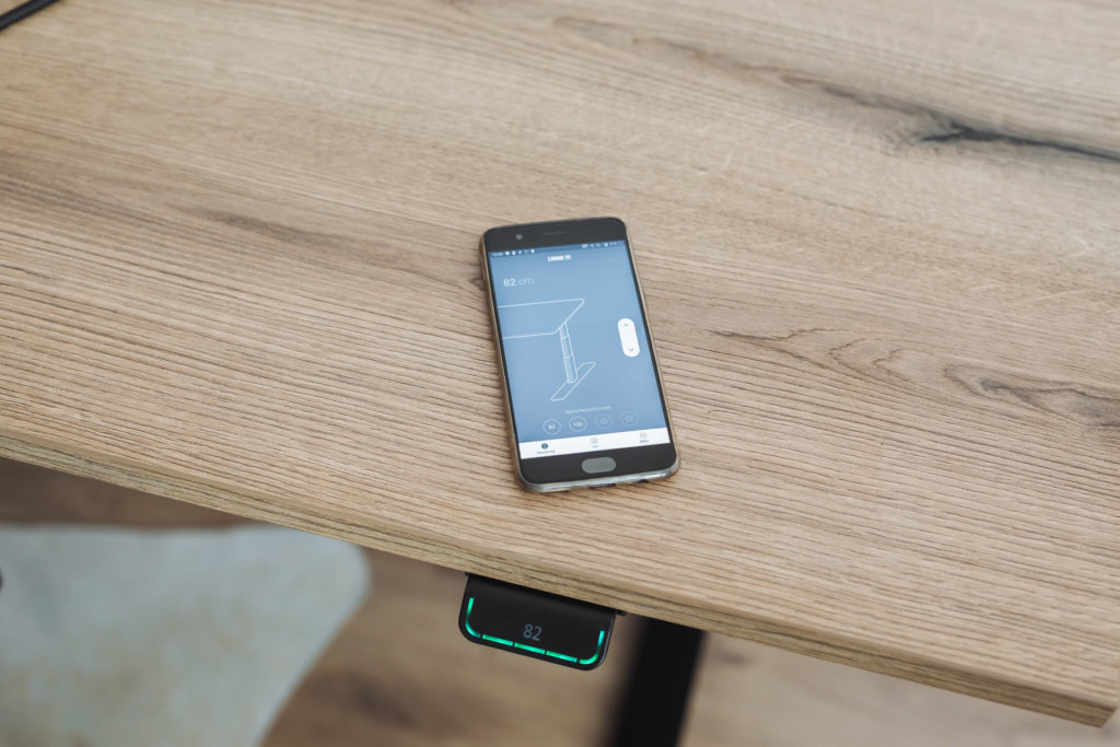 Detailaufnahme einer Tischplatte in Holzoptik, darauf ein Smartphone mit einer App zur Steuerung des Schreibtisches. Das digitale Bedienfeld unter der Tischplatte zeigt die eingestellte Höhe.