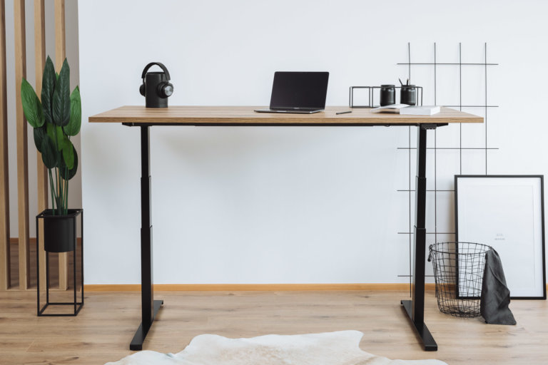 Ein höhenverstellbarer Schreibtisch mit schwarzem Gestell und einer Tischplatte in Holzoptik. Darauf ein Laptop, ein Bluetooth-Lautsprecher mit Kopfhörern sowie ein Notizbuch. Im Hintergrund eine Pflanze, ein Drahtkorb und eine minimalistische Wanddekoration.