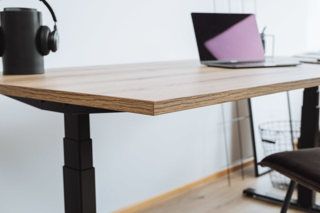 Ein höhenverstellbarer Schreibtisch mit Tischplatte in Holzoptik und schwarzem Gestell. Auf dem Tisch ein Laptop und ein Bluetooth-Lautsprecher mit Kopfhörern, ergänzt durch minimalistisches Zubehör.