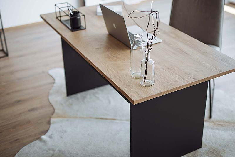 Schreibtisch mit der Oberfläche 'Eiche Sanremo', die eine natürliche Holzmaserung zeigt. Auf dem Tisch stehen zwei Glasvasen mit minimalistischen Ästen und ein Laptop. Der Tisch hat ein modernes Design mit dunklen Seiten und einem hellen Holzton, das auf einem Teppich mit weicher Struktur platziert ist.
