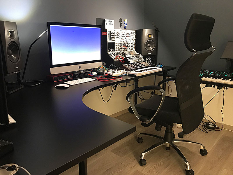 Professioneller Arbeitsplatz für Musikproduktion mit einer großen Tischplatte in der Oberfläche 'Schwarz FH'. Der Tisch bietet Platz für einen iMac, Studiomonitore, ein Keyboard, Synthesizer und eine modulare Synthesizer-Einheit. Ein ergonomischer Bürostuhl mit hoher Rückenlehne ergänzt die Arbeitsstation. Die Kabelmanagement-Elemente sind dezent unter der Tischplatte angebracht, während die Studiomonitore und Geräte optimal angeordnet sind.