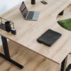 Eckschreibtisch mit Tischplatte in L-Form und Holzstruktur. Auf dem Eckschreibtisch steht ein grauer Laptop, eine Pflanze und Dekoration.