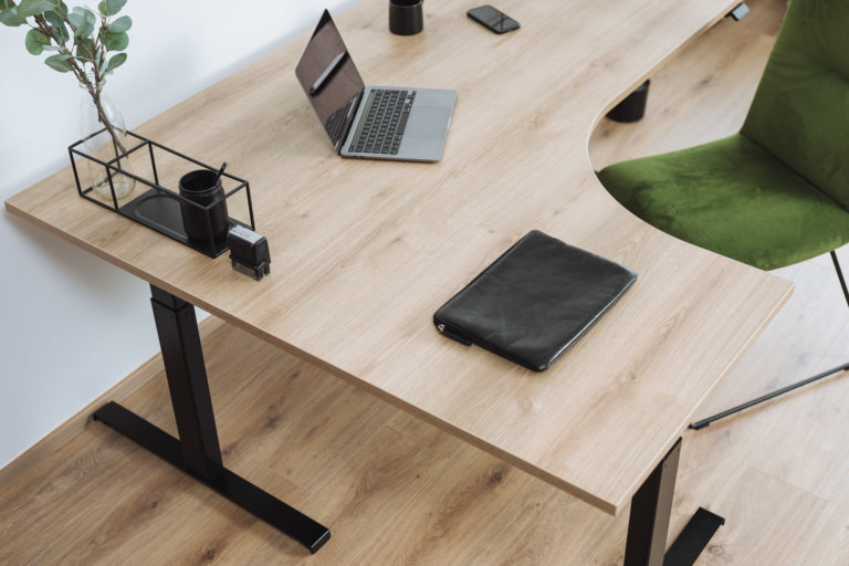 Eckschreibtisch mit Tischplatte in L-Form und Holzstruktur. Auf dem Eckschreibtisch steht ein grauer Laptop, eine Pflanze und Dekoration.