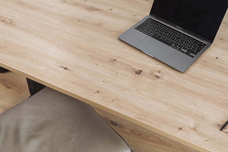 Tischplatte mit natürlicher Maserung und Astlöchern in der Oberfläche 'Atram Eiche'. Auf der Platte liegt ein Laptop. Der warme Farbton und die Struktur verleihen dem Tisch eine rustikale Eleganz.