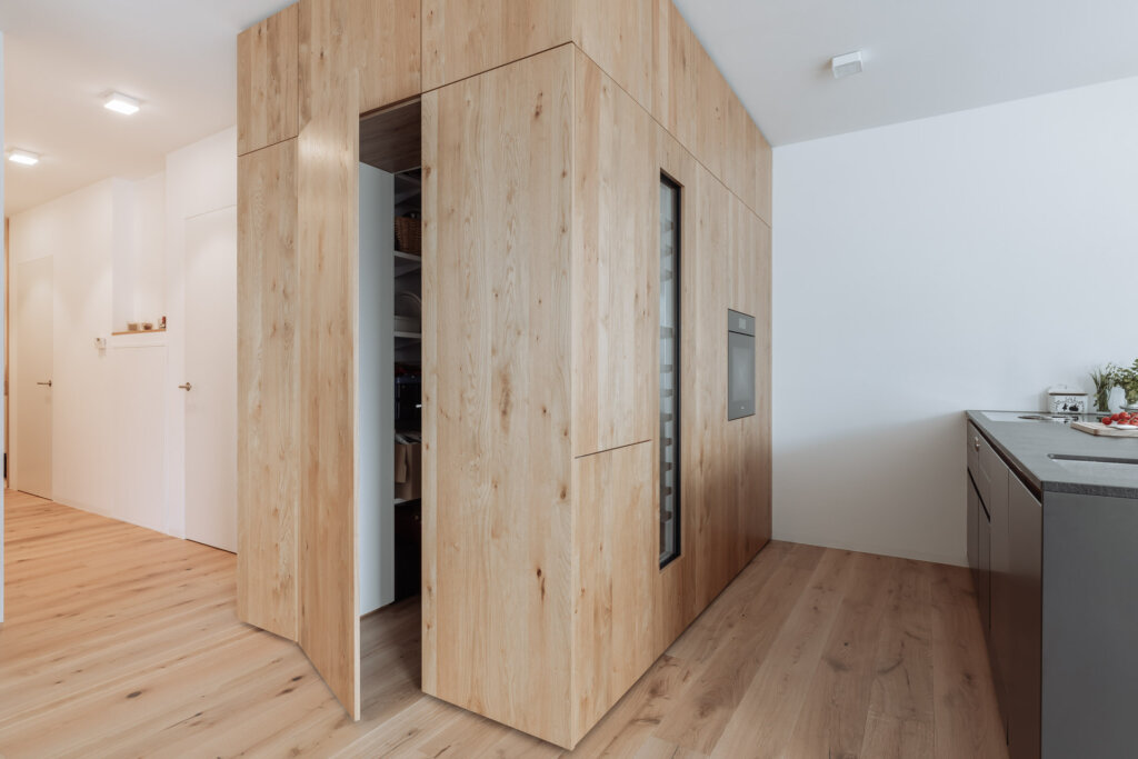 Moderne Küche mit einer Wand aus Schränken nach Maß in der Oberfläche 'Wildeiche furniert'. Die Schrankwand integriert eine Tür, die nahtlos in das Design übergeht, sowie einen Backofen und einen Weinkühlschrank. Rechts ist eine dunkle Küchenzeile mit einer Arbeitsfläche aus Stein zu sehen, während links der helle Flur mit minimalistischen weißen Türen anschließt.