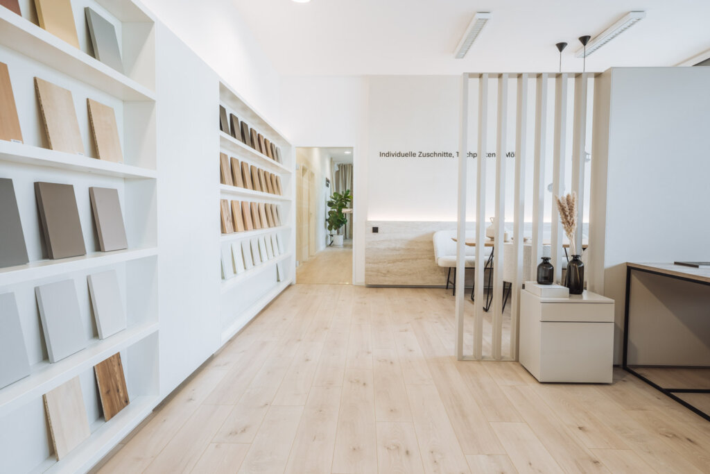 Moderner Schauraum mit hellen Holzfußböden und minimalistischen weißen Wänden. Auf Regalen entlang der linken Wand sind verschiedene Oberflächenmuster in Holz- und Neutralfarben ausgestellt. Im Hintergrund ist ein stilvoller Sitzbereich mit einer Trennwand aus vertikalen Holzstreben zu sehen, dekoriert mit schwarzen Vasen und Pampasgras.