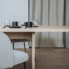 Ein moderner Esstisch aus hellem Holz, gedeckt mit minimalistischen schwarzen Tassen, Gläsern und einem schwarzen Teller. Im Vordergrund ein weißer Stuhl mit Stoffbezug, im Hintergrund dezente beige Vorhänge.