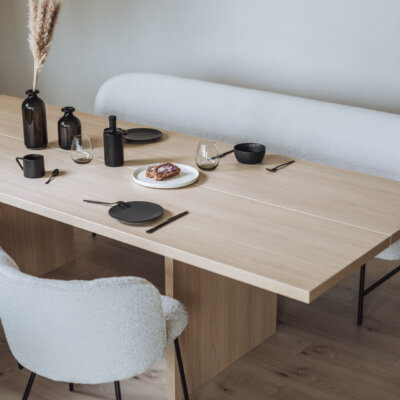 Ein moderner Essbereich mit einem hellen Holztisch, der mit minimalistischem schwarzen Geschirr, Gläsern und dekorativen schwarzen Vasen gedeckt ist. Im Hintergrund steht eine gemütliche weiße Polsterbank, ergänzt durch passende weiße Stühle mit schwarzem Gestell.