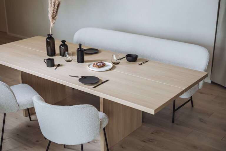 Ein moderner Essbereich mit einem hellen Holztisch, der mit minimalistischem schwarzen Geschirr, Gläsern und dekorativen schwarzen Vasen gedeckt ist. Im Hintergrund steht eine gemütliche weiße Polsterbank, ergänzt durch passende weiße Stühle mit schwarzem Gestell.