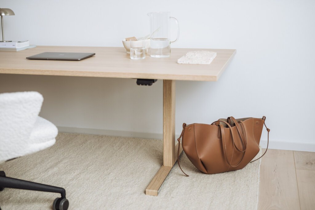 Detailansicht eines Tischbeins in Holzoptik, neben einer braunen Tasche auf einem Teppich.